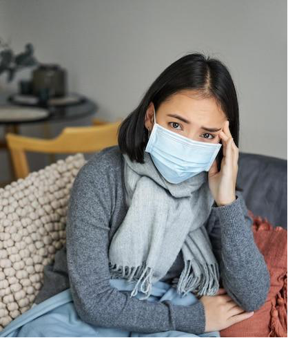 A young Asian woman is wearing a medical face mask and feeling unwell due to flu symptoms.